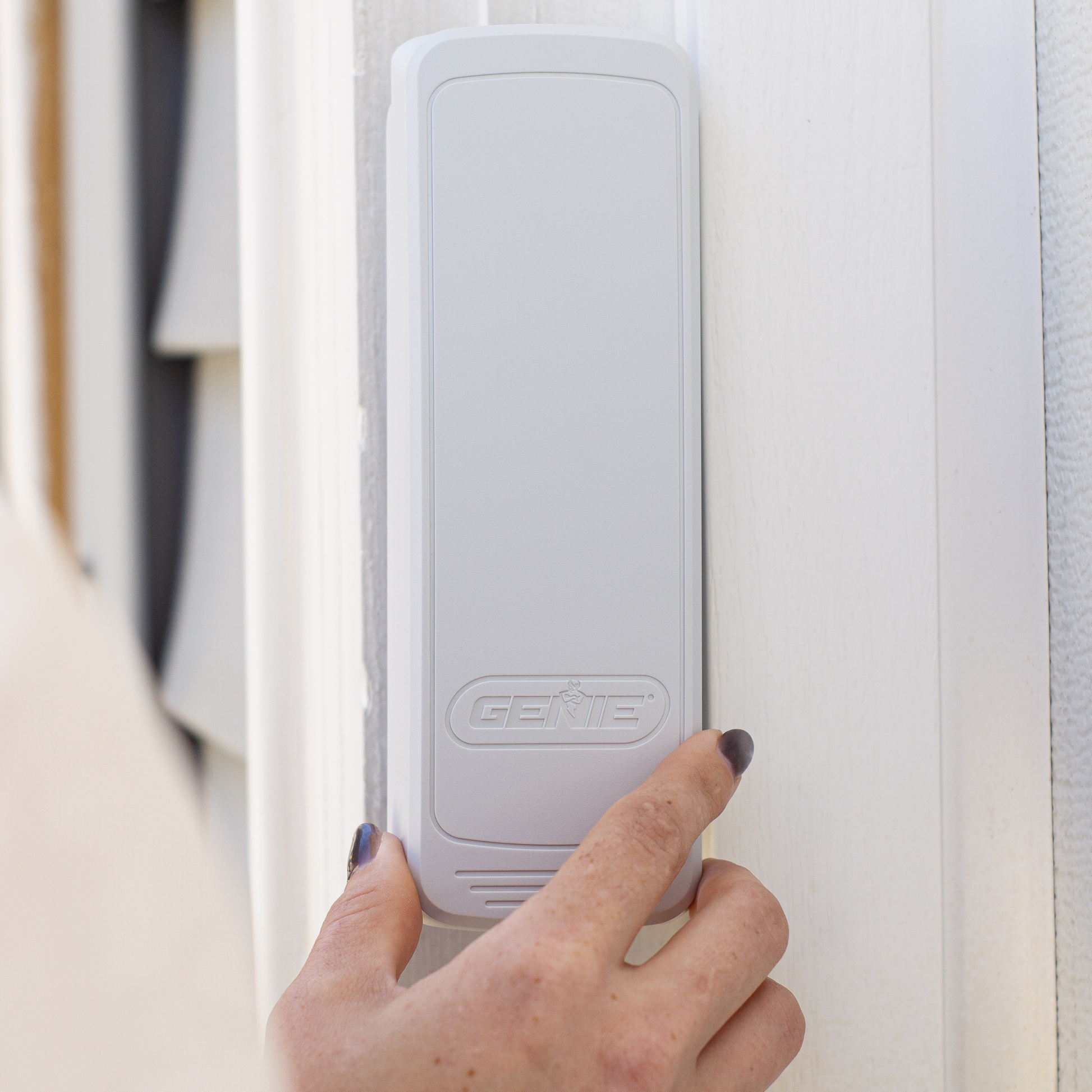 A hand with dark nail polish presses a button on a white rectangular Genie smart home device mounted on a white door frame.