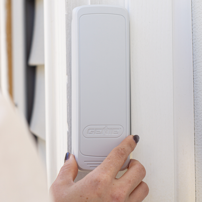 A hand with dark nail polish presses a button on a white rectangular Genie smart home device mounted on a white door frame.
